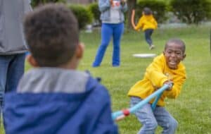 Two children competing in a tug-of-war on grass, with one smiling broadly while pulling the rope.
