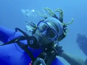 A diver with dreadlocks wears a mask and snorkel, surrounded by bubbles in deep blue water.
