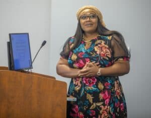 Bright African woman speaking at a conference, wearing a colorful floral dress and headwrap, standin.