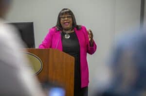 A woman in a pink blazer speaks passionately at a podium, gesturing expressively to the audience.