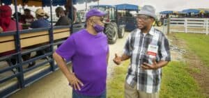 Two men engage in conversation outdoors at a community event, with a tractor and gravel path in the background.