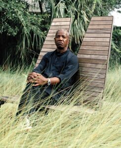 A man in a black outfit sits calmly on a wooden chair, surrounded by tall grass and greenery.