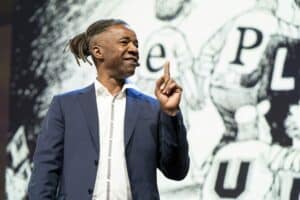 A man in a suit gestures confidently while speaking on stage, with a graphic backdrop.