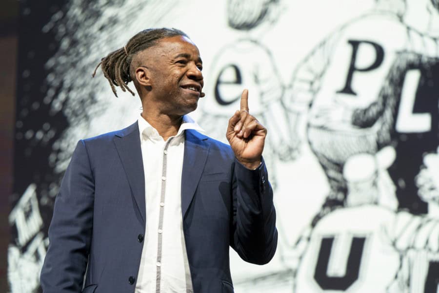 A man in a suit gestures confidently while speaking on stage, with a graphic backdrop.