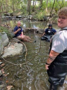 Group of people enjoying a stream in a forested area, taking photos and relaxing outdoors.