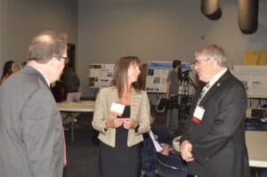 Three professionals engage in conversation at a conference, with posters and a camera crew in the background.