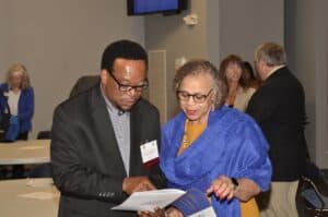 Two people discuss a document, one wearing glasses and a blue shawl, in a room with several attendees.