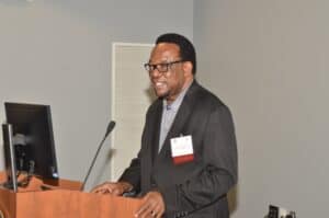 A speaker presenting at a podium with a microphone, wearing glasses and a blazer.