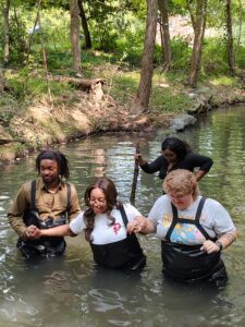 Group of diverse people wading in a river surrounded by trees during outdoor activity.