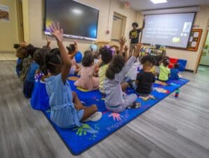 Kids in a classroom raising their hands during a lesson with a teacher and a digital screen.