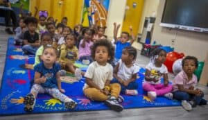 A diverse group of young children sitting on a colorful rug, engaged and listening attentively in a classroom setting.