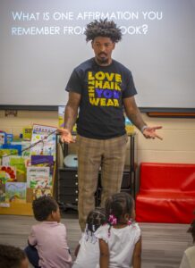 A speaker engages a group of children, discussing affirmations in a classroom setting. Books are displayed in the…