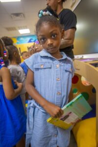 A young girl in a denim dress holds a book, looking thoughtfully at the camera amidst other children.
