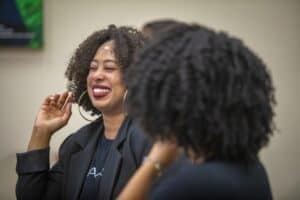 A woman with curly hair smiles broadly, laughing while engaging with others in a lively conversation.