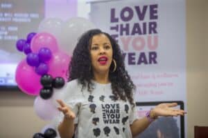 A woman with curly hair speaks passionately while wearing a t-shirt that promotes natural hair appreciation.