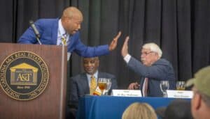 High-five between two men at a formal event with a panel and audience, promoting real estate and pro.