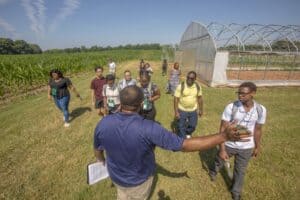 A group of students listens to an instructor inside a greenhouse. The instructor, wearing a blue shirt, gestures while holding a clipboard, explaining rows of small plants growing in the soil. The students, wearing backpacks, stand at the entrance, observing the setup. The greenhouse structure is made of clear panels, allowing sunlight to illuminate the interior. Outside, green grass is visible.