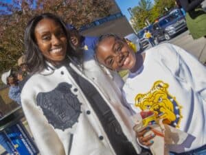 Two smiling women wearing CAES News branded apparel outdoors on a sunny day.