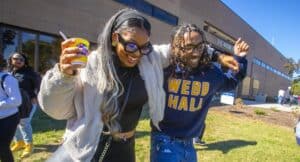 Jahbez Roach, wearing a sweatshirt that reads 'Webb Hall,' dances joyfully with a friend holding a cup, at the College of Agriculture and Environmental Sciences (CAES) Annual Alumni Homecoming Celebration and Cookout on the lawn at B.C. Webb Hall. The event included games, food, fellowship, entertainment, and Aggie spirit.