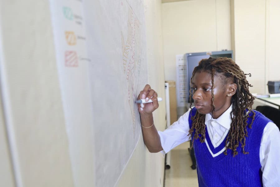 Jaliyah Shepherd, a sophomore landscape architecture major, works intently on a site plan, drawing with a marker on a large paper on the wall. She wears a blue sweater vest over a white shirt. Part of the Landscape Architecture Foundation Ignite Scholar program, she receives scholarships, internships, and mentorship opportunities.