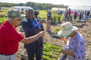 Dr. Jimo Ibrahim, an Agricultural Health and Safety Specialist from N.C. A&T State University Cooperative Extension, speaks with Luciano and Maria Alvarado in a community garden during the Regional Small Farms Conference on October 7-8, 2024, at N.C. A&T University Farm Pavilion. Several participants are visible in the background, exploring the garden beds and plants