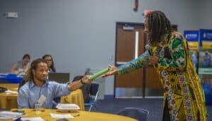 A woman in colorful traditional attire hands a green object to a seated woman at a conference table.
