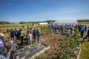 Community gathering at a farm with greenhouse and vegetable garden under clear blue sky.