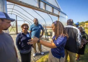 Group of people engaged in discussion near a greenhouse on a sunny day.