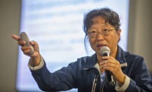 Dr. Kathleen Liang, conference organizer, speaks into a microphone while holding a presentation remote. She gestures with her hand, emphasizing a point during her talk at the Regional Small Farms Conference. A projected slide is blurred in the background.