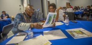 Two people share and discuss printed images at a table in a community event. Various materials are visible around them.