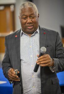 A man in a checked blazer holds a microphone, engaged in conversation. He appears confident and approachable.