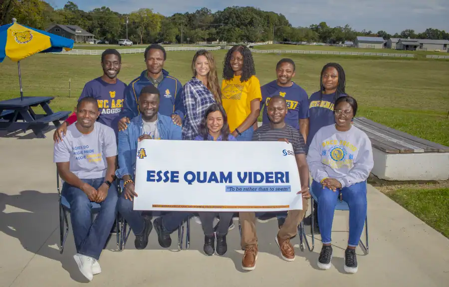 Group photo of the team Esse Quam Videri, which translates to 'To be rather than to seem,' holding a banner with their team name. The group of eleven people smiles at the camera, with a grassy field and farm buildings in the background. Some team members wear North Carolina A&T State University apparel.