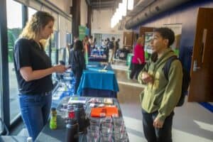 A career fair in a bright, modern hallway with tables covered in blue and green cloths. On the left, a representative wearing a black shirt and jeans speaks with a student in a green hoodie and backpack. The tables display promotional items such as water bottles, brochures, and cups. Other attendees and representatives are visible in the background, engaging in conversations.