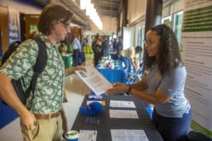 A student wearing a green, tropical-print shirt and backpack hands a resume to a representative in a blue, striped polo shirt at a career fair. They are standing at a table covered in informational materials, including brochures, pens, and a blue hat. In the background, other attendees and representatives interact at different booths.