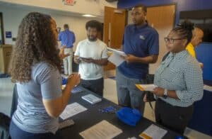A representative in a blue striped shirt speaks to three students gathered around a table during a career fair. The students hold brochures and notepads, listening attentively. The table displays informational materials, pens, and a blue hat. In the background, additional attendees and representatives engage in discussions.