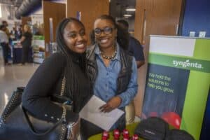 Two women smiling at an event booth. One is wearing a black sweater and carrying a black handbag, while the other wears glasses, a denim shirt, and a black vest, holding a paper. The booth displays a green Syngenta banner with the slogan "We hold the solutions to help you grow." Small model tractors and branded hats are on the table. Other attendees and booths are visible in the background.