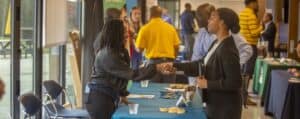 A student shakes hands with a representative at a career fair booth. The booth has a blue tablecloth with pens, pamphlets, and cookies displayed. Other attendees and representatives engage in conversations at nearby booths. The event is set in a bright space with large windows and various informational displays in the background.