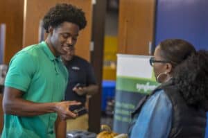 A young man in a green polo shirt smiles while holding a smartphone, engaging with a woman wearing glasses and a black vest. They appear to be at an indoor event, with a blurred banner and another person in the background.