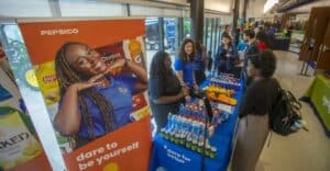 A PepsiCo booth at an indoor career fair features multiple representatives engaging with a student. The booth displays a variety of snacks and beverages, including chips and bottled drinks. Prominent branding and banners with slogans such as "dare to be yourself" and "dare for better" are visible. The background shows other booths and attendees interacting.
