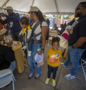 A family enjoys Ag & Environmental Science Day under a large event tent. A mother holds a baby in a carrier, while a young girl in a bright yellow hoodie smiles widely. People are seated at tables in the background, eating and talking, creating a lively and cheerful atmosphere.