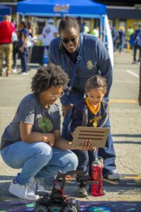 An adult and a young child interact with a tablet while observing a small robot on a mat during Ag & Environmental Science Day. Another adult stands nearby, smiling as they watch. The event takes place outdoors with tents and other attendees visible in the background.