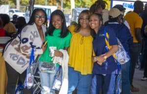 Four women smile and pose together under a tent during Ag & Environmental Science Day. They are holding event bags and enjoying the lively atmosphere, with other attendees and activities visible in the background."