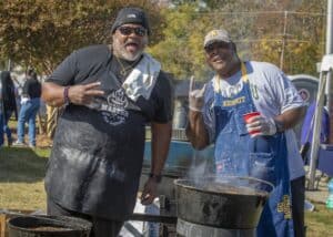 Two men, wearing aprons with 'Grill Master' and 'Go Aggies' respectively, pose with smiles and hand gestures in front of a grill during Ag & Environmental Science Day. Steam rises from the cooking pots, and the event's lively atmosphere is visible in the background.