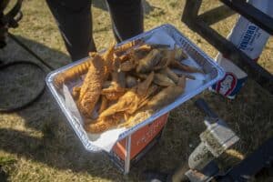 A metal tray filled with freshly fried fish, resting on a box labeled "frying oil," outdoors on a grassy surface. A bag of charcoal is partially visible in the background.