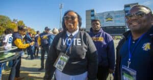 A group of people gathered outside Truist Stadium on a sunny day. In the foreground, a smiling woman wearing a "VOTE" sweatshirt and sunglasses is accompanied by others wearing N.C. A&T Aggies apparel. Lanyards with event badges are visible, indicating a university event.