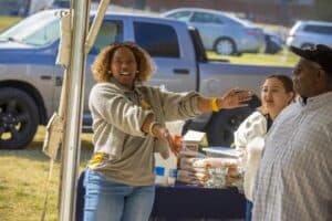 A woman wearing an N.C. A&T shirt gestures enthusiastically under a tent, while two other individuals stand nearby. The table behind them holds food items, including bread and snacks. A parked truck is visible in the background.