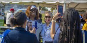 Two women pose for a photo under a tent at an outdoor event. One wears an N.C. A&T lanyard and sunglasses, while the other smiles in a blue N.C. A&T shirt. Several people in the background use their phones to capture the moment.