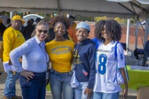 Four people, including a woman in a blue N.C. A&T shirt and three young adults in N.C. A&T apparel, smile and pose together under a tent at an outdoor event. Other attendees and tables with green tablecloths are visible in the background.