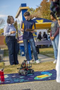 A young man holding a clipboard explains a robotics demonstration to an older woman and another person standing nearby at an outdoor event. A small robotic vehicle sits on a colorful space-themed mat on the ground. A tent and event signage are visible in the background.
