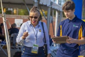A woman wearing an N.C. A&T shirt and lanyard smiles and gestures enthusiastically while speaking with a young man holding a tablet. They are outdoors under a tent at an event, with other attendees visible in the background.